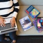 Woman using laptop on sofa, surrounded by programming books, learning coding.
